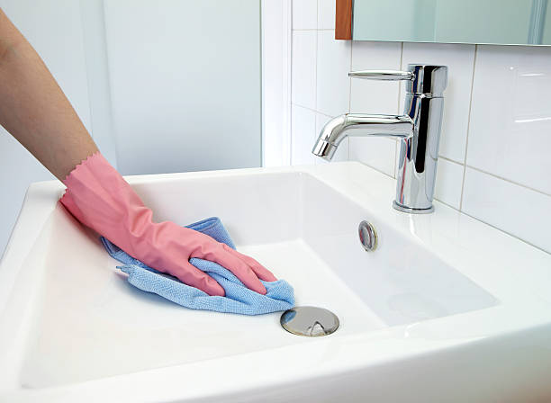 A person wearing pink gloves is cleaning a white bathroom sink with a blue cloth.